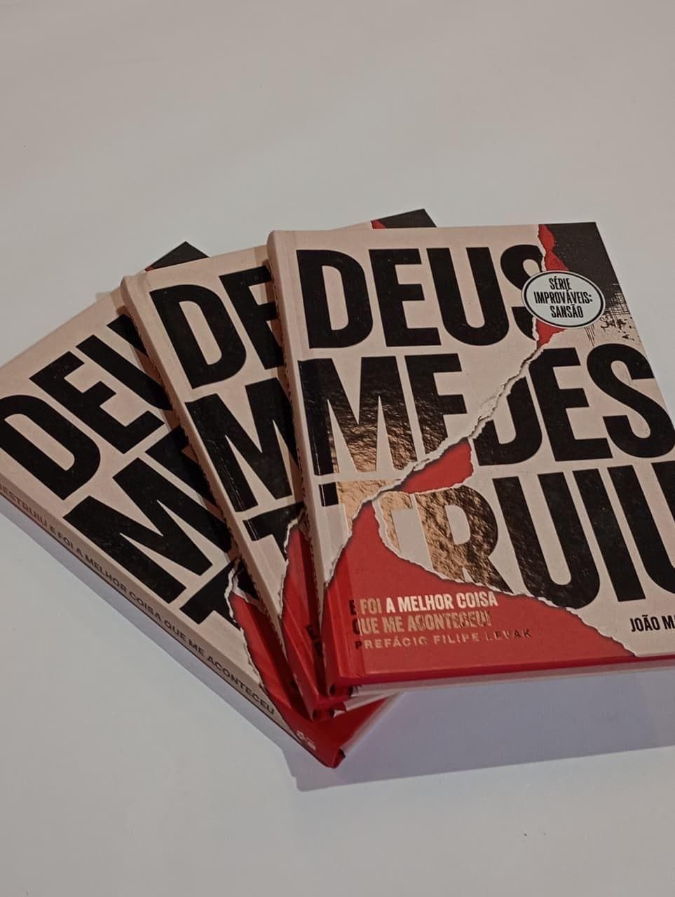 Três exemplares de Deus me destruiu abertos em leque, mostrando a capa vermelha e creme